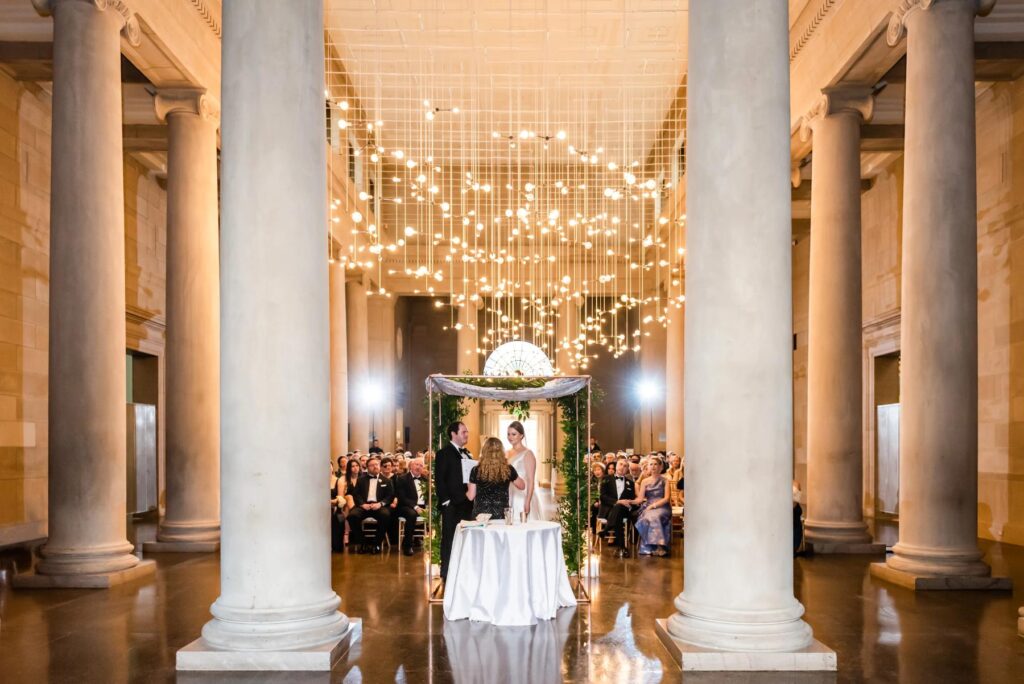 A wedding ceremony with a bridee and groom with lights hanging down at a Baltimore Museum of Art Wedding planned by Blue Sapphire Events.